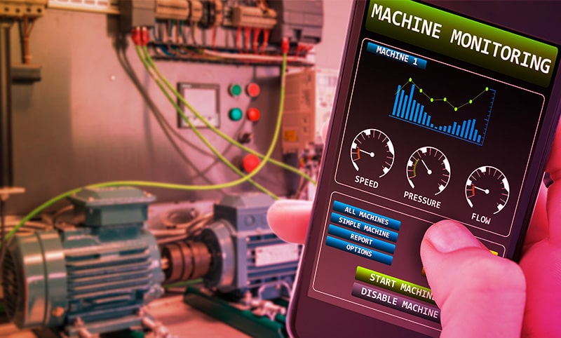 A hand in the foreground holding a smart phone with a smart monitoring app displaying. Industrial motors are in the background.