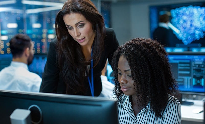 A woman engineer looking at a computer with another woman standing over her showing her something on the screen